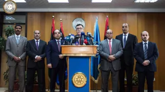 The image shows a group of eight men standing in a formal setting, likely a press conference or official announcement. There is a podium at the center with a seal or emblem, and the speaker appears to be addressing the audience. The backdrop features flags, suggesting a governmental or institutional context. The men are dressed in suits and ties, conveying a professional atmosphere.