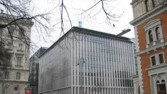 این تصویر نمای یک ساختمان مدرن را نشان می‌دهد که دارای طراحی مینیمالیستی است. نمای ساختمان با پنجره‌های عمودی و خطوط باریک تقسیم شده، ظاهری مرتب و شیک دارد. اطراف ساختمان ممکن است شامل خودروها و درختان بدون برگ در فصل زمستان باشد. در سمت چپ و راست تصویر، ساختمان‌های دیگری با طراحی‌های کلاسیک‌تر وجود دارند که تضادی جالب با ساختمان مدرن ایجاد می‌کنند. فضا به نظر می‌رسد که در یک منطقه شهری و ممکن است در فصل سرد سال باشد.