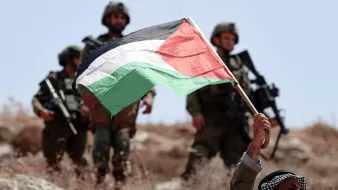 L'immagine mostra un uomo che regge alta una bandiera palestinese, composta dai colori nero, bianco, verde e rosso, mentre si trova su un terreno roccioso. Dietro di lui, si possono vedere soldati armati, presumibilmente appartenenti a una forze militari, che osservano la scena. L'atmosfera è tesa, e il contesto sembra essere legato a un conflitto.