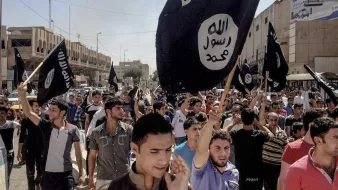 L'image montre une foule de personnes rassemblées dans une rue, tenant des drapeaux noirs emblématiques. Les manifestants semblent mobilisés et expriment une forte émotion. L'environnement urbain autour d'eux montre des bâtiments et une rue animée, suggérant un événement significatif ou une manifestation.