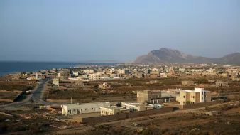 تظهر الصورة منظرًا خلابًا لبلدة ساحلية تطل على البحر. يُظهر المشهد منازل مبنية بشكل متفرق، بعضها حديث والآخر تقليدي، مع شوارع تمتد بين المباني. في الخلفية، يمكن رؤية سلسلة جبال، بينما يمتد البحر الفسيح على الجهة اليسرى. السماء صافية وتبدو أشعة الشمس مشرقة، مما يضفي شعورًا بالهدوء والجمال الطبيعي على المكان.