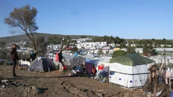 تصویر نشان‌دهنده یک منطقه با وضعیت نامناسب است که در آن چادرهایی به عنوان محل سکونت نصب شده‌اند. در پس‌زمینه، زمین‌های ناهموار و درختانی مشاهده می‌شود. پس‌زمینه شهری با ساختمان‌هایی دیده می‌شود که نسبتاً به چادرها نزدیک هستند. احساس کلی تصویر بیانگر شرایط سخت زندگی افرادی است که در این چادرها سکونت دارند. آسمان آبی و روز روشن است، اما وضعیت محیط نشان‌دهنده فقر و مشکلات معیشتی است.