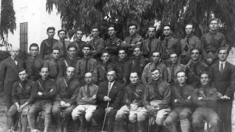 L'image montre un groupe de soldats en uniforme, probablement pris au début du 20e siècle. Ils sont assis et debout en rangée, avec des expressions sérieuses. Au centre, un homme en costume, tenant une canne, semble superviser le groupe. L'arrière-plan présente des arbres feuillus, ajoutant une touche naturelle à la scène. L'ambiance générale évoque une certaine solennité et un sentiment de camaraderie parmi les membres du groupe.