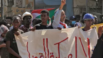 L'image montre une scène de manifestation, probablement en rapport avec des revendications politiques. On y voit des personnes tenant une grande bannière. Un homme au premier plan fait un signe de paix avec ses doigts. Les manifestants semblent déterminés et engagés, et des drapeaux sont visibles en arrière-plan, indiquant un contexte national. L'atmosphère est chargée d'émotion, illustrant un moment de solidarité.