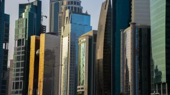 L'image montre une vue urbaine moderne avec plusieurs gratte-ciels aux façades brillantes et réfléchissantes. Les bâtiments sont de différentes formes et tailles, avec des matériaux en verre et en métal qui scintillent sous la lumière. On peut remarquer un mélange d'architectures contemporaines, certaines avec des courbes élégantes et d'autres plus angulaires. Le paysage urbain reflète un environnement dynamique et en pleine croissance, typique des grandes villes modernes.