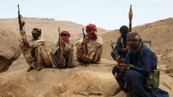 L'image montre un groupe de personnes assises sur des rochers dans un paysage désertique. Elles portent des vêtements traditionnels, masquent partiellement leur visage avec des foulards et tiennent des armes. L'environnement est montagneux et aride, avec des formations rocheuses en arrière-plan. L'ambiance semble sérieuse et reflète une situation de tension.