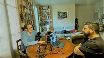 The image depicts two men engaged in a conversation at a round wooden dining table. One man, wearing a light blue sweater and glasses, appears to be animatedly speaking and gesturing, while the other man, dressed in a black shirt, is listening attentively. On the table, there are microphones and a laptop, suggesting they might be recording a podcast or conducting an interview. In the background, a cozy living space is visible with shelves full of books and a sofa where another person seems to be resting. The overall atmosphere feels informal and collaborative.