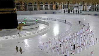 The image depicts a scene from the Grand Mosque in Mecca, known as the Masjid al-Haram. It features the Kaaba, a black cube-shaped structure covered in a black cloth with gold embroidery, which is the focal point of Islamic worship. Surrounding the Kaaba, there are congregational worshippers who appear to be lined up in rows, praying. The area is spacious and well-lit, with white marble flooring. In the background, there are architectural elements characteristic of the mosque, including arched structures. Security personnel can also be seen in the image, emphasizing the organized nature of the gathering.