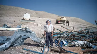 L'image montre un homme debout au milieu d'un terrain désolé. Il est entouré de débris, comprenant des structures métalliques et des morceaux de toile. En arrière-plan, on peut voir des réservoirs ou des citernes, ainsi qu'un cheval. Le paysage semble aride, avec des collines en arrière-plan sous un ciel bleu. L'homme porte une tenue simple et dégage une impression de résilience dans un environnement difficile.