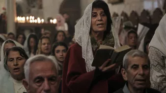 L'immagine mostra un gruppo di persone durante una cerimonia, probabilmente in una chiesa. Una donna al centro, che indossa un velo bianco e un abito rosso, tiene in mano un libro e sembra partecipare attivamente alla celebrazione. Intorno a lei ci sono altre persone, alcune delle quali appaiono in preghiera o cantando. L'atmosfera è solenne, con un gioco di luci morbide e candele accese sullo sfondo che creano un'illuminazione calda e accogliente.