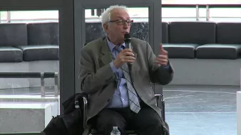 Il s'agit d'un homme assis sur une chaise, tenant un microphone, probablement en train de parler ou de donner une présentation. Il porte une veste et une cravate, et l'environnement semble être un espace public ou une salle de conférence. Des éléments modernes de design sont visibles en arrière-plan.