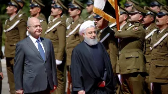 در این تصویر، دو شخصیت در حال برگزاری یک مراسم رسمی هستند. در سمت چپ، فردی با لباس رسمی و کت و شلوار ایستاده است. در سمت راست، فرد دیگری با لباس سنتی ایرانی، که به یک عمامه و عبای سیاه پوشیده است، حضور دارد. در پس‌زمینه، تعدادی از نظامیان لباس‌پوشیده به خط ایستاده‌اند و در حال انجام تشریفات نظامی هستند. تصویر نشان‌دهنده یک رویداد رسمی یا دیدار دیپلماتیک است.