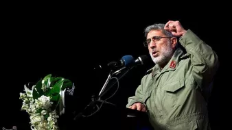 در این تصویر، یک فرد در حال سخنرانی در یک مکان رسمی است. او لباس نظامی به تن دارد و در حال بیان نکاتی با حرکات دست است. در کنار او، میزی با گل‌های تزئینی قرار دارد و میکروفون و بلندگو نیز دیده می‌شود. پس‌زمینه تصویر تاریک است و تمرکز بیشتری بر روی سخنران وجود دارد.