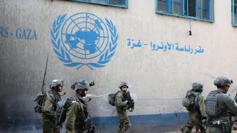 تظهر الصورة مجموعة من الجنود يرتدون زيًا عسكريًا ويقومون بالتحرك في منطقة قريبة من مبنى يحمل شعار الأمم المتحدة باللغة العربية. الجدران تحمل كتابة تشير إلى مقر الأونروا في غزة، ويبدو أن المكان يعاني من بعض الفوضى أو الأضرار. الجنود يظهرون وكأنهم في حالة تأهب، مما يعكس أجواء من التوتر أو النشاط العسكري في المنطقة.