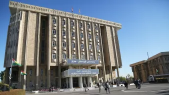 تظهر الصورة مبنى البرلمان في إقليم كردستان العراق. المبنى يتميز بتصميمه المعماري العصري، ويتكون من عدة طوابق مع واجهة تحمل لافتة كبيرة تحمل اسم البرلمان. في المقدمة، يمكن رؤية أشخاص يمشون حول المبنى، بالإضافة إلى أعلام كردية ترفرف في المكان. السماء زرقاء وتضفي جواً من النشاط والحيوية على الصورة.