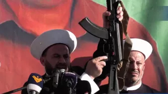 The image depicts two men, wearing white turbans and traditional garments, standing at a podium with microphones. One man is prominently holding an assault rifle above his head, while the other looks on. The background features a large, colorful mural or poster, possibly of a political figure or symbol. The scene suggests a demonstration or rally, conveying themes of power and assertiveness.