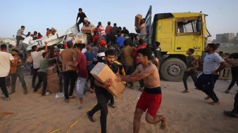 L'immagine mostra un gran numero di persone, principalmente giovani uomini, che sono radunati attorno a un camion giallo. Alcuni di loro sembrano scaricare scatole, mentre altri stanno trasportando i pacchi. L'ambiente appare polveroso e le persone sono visibilmente attive e impegnate nell'operazione di distribuzione, con espressioni di urgenza. Alcuni individui sono a torso nudo e indossano abiti vari. Si percepisce un'atmosfera di collaborazione nel tentativo di ricevere e distribuire i rifornimenti.