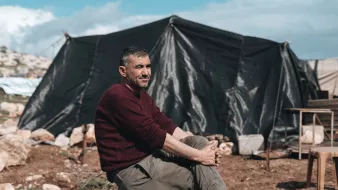 Nell'immagine si vede un uomo seduto su una roccia in un ambiente all'aperto, con un'espressione rilassata. Indossa un maglione scuro e pantaloni chiari. Dietro di lui si nota un grande tendone nero che fa da rifugio, immerso in un paesaggio di terra e rocce. Il cielo è parzialmente nuvoloso, dando un'atmosfera di calma all'ambiente circostante.