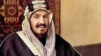 L'image montre un homme assis, portant un habit traditionnel. Il est vêtu d'une thobe sombre et d'un ghutrah, un couvre-chef caractéristique de la culture arabe. Il tient une canne et affiche une expression calme et posée. L'arrière-plan présente un motif décoratif qui renforce le contexte culturel de la scène. Les couleurs de l'image sont enrichies, donnant vie aux vêtements et à l'environnement.