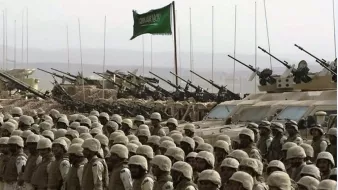 تظهر في الصورة مجموعة من الجنود يرتدون زيًا عسكريًا واقفين في صفوف منتظمة. خلفهم، توجد دبابات ومدرعات عسكرية، حيث تُشاهد المدافع موجهة نحو الأمام. في خلفية الصورة، يوجد علم أخضر يرفرف، مما يوحي بأن هذه الصورة مرتبطة بقوة عسكرية أو استعراض. تعكس الصورة أجواءً عسكرية وهيبة القوات المدافعة.