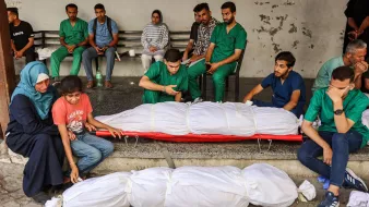 L'immagine mostra un gruppo di persone in uno spazio pubblico. Alcuni individui indossano camici verdi, suggerendo che potrebbero essere operatori sanitari. In primo piano, ci sono due corpi avvolti in lenzuola bianche. Alcuni membri del gruppo sembrano essere in uno stato di lutto, mentre altri osservano la scena in modo serio. L'atmosfera complessiva è di tristezza e riflessione.
