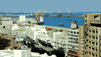 L'image montre un paysage urbain avec des bâtiments blancs et modernes. On aperçoit une vue sur le port, avec des navires et des grues au loin. Le ciel est clair, ce qui laisse transparaître une ambiance paisible. Les structures architecturales semblent principalement de couleur claire, créant un contraste avec la mer bleue. C'est une scène qui évoque l'activité maritime et la vie urbaine.
