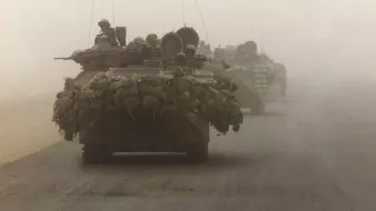 La imagen muestra un convoy militar en un ambiente polvoriento y desértico. Se pueden ver varios vehículos blindados, posiblemente tanques, avanzando en formación. Los vehículos están cargados con equipos y suministros, lo que sugiere que están en una misión de combate o de suministro. La visibilidad es limitada debido a la niebla o polvo, lo que añade una atmósfera tensa a la escena.