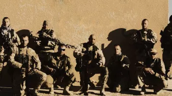 الصورة تظهر مجموعة من الجنود أو أفراد القوات المسلحة، يقفون معًا في وضعية تعكس القوة والانضباط. يرتدون زيًا عسكريًا ويحملون أسلحة، ويظهرون بمظهر جاد. الخلفية تبدو بسيطة، مما يركز الانتباه على المجموعة. تعكس الصورة روح الفريق والتضامن بين الأفراد.