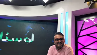L'immagine mostra un uomo sorridente seduto davanti a una scrivania in uno studio televisivo. Indossa una camicia rosa e presenta un aspetto rilassato e accogliente. Dietro di lui si possono notare schermi che mostrano un logo o un testo in arabo, mentre l'illuminazione dello studio è accentuata da luci colorate, in particolare tonalità di viola e blu. L'ambiente sembra moderno e professionale, tipico di una trasmissione televisiva.