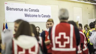 L'image montre un intérieur où un groupe de personnes se rassemble. Au fond, il y a une grande affiche indiquant "Bienvenue au Canada" en français et en anglais. Plusieurs personnes portent des vestes rouges avec une croix blanche, ce qui suggère qu'elles sont des secouristes ou des membres d'une organisation humanitaire. L'ambiance semble accueillante et axée sur l'accueil des nouveaux arrivants. Des drapeaux canadiens sont également visibles dans la scène.