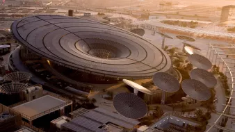 L'image montre un bâtiment architectural moderne et futuriste, avec un design en forme d'anneau ou de soucoupe volante. Le toit est en grande partie vitré, permettant à la lumière naturelle d'entrer. Des structures circulaires, semblables à des panneaux solaires ou des œuvres d'art, entourent le bâtiment, ajoutant à son aspect innovant. Le paysage environnant semble désertique, avec des infrastructures modernes visibles à l'arrière-plan, indiquant un environnement urbain en développement. La lumière douce du coucher du soleil donne une ambiance chaleureuse à la scène.