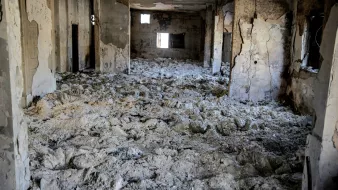 L'immagine mostra un interno di un edificio gravemente danneggiato. Le pareti sono deteriorate e presentano crepe, mentre il soffitto appare scrostato. Il pavimento è coperto da una massa bianca, che potrebbe essere polvere o detriti. I fori nelle pareti e nelle finestre suggeriscono che l'area è stata abbandonata e distrutta, creando un'atmosfera di desolazione e degrado. La luce che entra attraverso le aperture crea un contrasto con le ombre all'interno dello spazio.