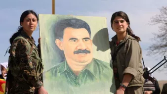L'image montre deux femmes en tenue militaire tenant un portrait d'un homme avec une moustache. Elles semblent participer à un événement ou une manifestation, entourées d'une atmosphère qui indique une forme de dévotion ou de commémoration. Le fond est naturel, avec un ciel partiellement nuageux. Les expressions des femmes suggèrent détermination et fierté.