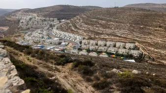 L'immagine mostra un paesaggio collinare, caratterizzato da una zona abitata con edifici bianchi disposti lungo una strada che si snoda tra le colline. In primo piano, si vede una vegetazione bassa e cespugliosa. Sullo sfondo, le colline presentano strati di roccia esposti, creando un effetto di stratificazione geologica. La luce appare naturale, suggerendo un'ora del giorno in cui il sole è alto, illuminando la scena con tonalità calde.