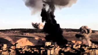 L'image montre une scène de destruction, probablement liée à un conflit armé. On aperçoit des colonnes de fumée noire s'élevant dans le ciel, suggérant des explosions récentes. En arrière-plan, on voit des collines et des ruines de bâtiments, ce qui témoigne des effets dévastateurs sur l'environnement urbain. Le paysage semble désolé, évoquant une atmosphère de chaos et de désespoir.