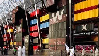The image depicts a modern architectural structure that resembles a stadium or a large public venue. The building features a striking façade made up of brightly colored shipping containers in red, yellow, and blue, arranged in a visually dynamic way. There are tall, angular elements with a white roof framework above. In the foreground, a group of people dressed in traditional attire can be seen, indicating a cultural context. Overall, the design conveys a sense of contemporary style and innovative use of space.