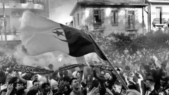 L'image montre une scène vibrante et animée, probablement lors d'un événement festif ou d'une manifestation. On peut voir une grande foule de personnes rassemblées, certaines brandissant des drapeaux et des écharpes. Les visages expriment la joie et l'enthousiasme. Une atmosphère de célébration est accentuée par des fumigènes qui créent un léger brouillard. En arrière-plan, on distingue des bâtiments, ce qui suggère que cette scène se déroule en milieu urbain.