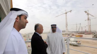 L'image montre trois hommes, apparemment en discussion, sur un balcon ou une plateforme. Ils sont vêtus de vêtements traditionnels des Émirats, comprenant des kandouras et des ghutras. En arrière-plan, on peut voir un site de construction avec des grues et des véhicules, suggérant un projet de développement en cours. L'environnement est poussiéreux et légèrement brumeux, typique des zones de construction dans des climats chauds. Les hommes semblent sérieux et engagés dans leur conversation.