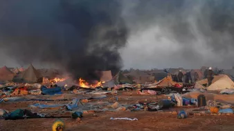 L'image montre un camp de tentes dans un environnement chaotique. On peut voir de la fumée noire s'élever dans le ciel, accompagnée de flammes, indiquant un incendie. Le sol est encombré de débris et de couvertures abandonnées, ce qui suggère une situation difficile. En arrière-plan, des personnes sont visibles, probablement des secouristes ou des autorités, tentant de gérer la situation. L'atmosphère est sombre et perturbée, reflétant une scène de crise ou de catastrophe.