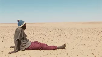 L'image montre un homme assis sur le sol désertique. Il porte un pull avec un bonnet et un pantalon de couleur prune. Le paysage autour de lui est vaste et aride, constitué principalement de sable et de petites pierres, s'étendant à perte de vue sous un ciel clair. L'atmosphère semble à la fois isolée et silencieuse, évoquant une sensation de solitude dans cet environnement désertique.