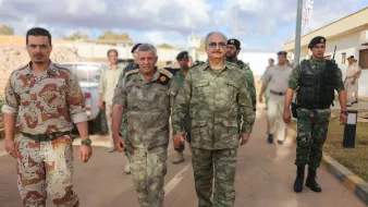 L'image montre un groupe de militaires marchant ensemble dans un environnement urbain. Deux hommes en avant-plan semblent discuter tout en marchant côte à côte. L'un d'eux porte un uniforme camouflage de couleur verte, tandis que l'autre a un uniforme militaire gris avec des distinctions. En arrière-plan, on peut voir d'autres soldats et des véhicules militaires, ce qui indique qu'ils se trouvent dans une zone potentiellement sécurisée. Les soldats semblent concentrés et en mission.