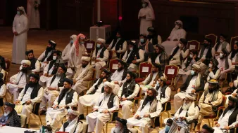 تظهر الصورة مجموعة من الأشخاص يجلسون في قاعة كبيرة، يرتدون ملابس تقليدية. يبدو أن بعضهم يرتدي الأشرطة والعمائم، بينما يحضر آخرون بزي رسمي. القاعة مزينة بروعة، وهناك تفاصيل معمارية خلفهم، مما يشير إلى أنها حدث رسمي أو مؤتمر. يبدو أن بعض الحضور يرتدون كمامات، مما يدل على الاحتياطات الصحية.