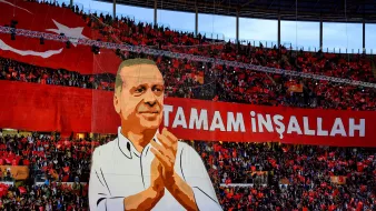 The image depicts a large crowd in a stadium, with a significant banner displayed prominently. The banner features a representation of a person, possibly a political figure, who is shown applauding. Surrounding the figure are enthusiastic supporters holding a sea of red flags or banners. The text "TAMAM İNŞALLAH" is written in bold letters, which translates to "It will be done, God willing" in English, conveying a message of hope or determination. The overall atmosphere appears vibrant and charged with energy, indicative of a political rally or celebration.
