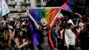 L'immagine mostra un gruppo di persone in una strada affollata, probabilmente durante una manifestazione o una parata. Al centro, una giovane donna con capelli corti e bianchi è in primo piano, mentre tiene alta una grande bandiera arcobaleno. Sembra esprimere gioia e determinazione. Intorno a lei, altre persone sventolano bandiere e manifestano il loro sostegno, creando un'atmosfera di festeggiamento e orgoglio. Le decorazioni e i colori vivaci trasmettono un messaggio di inclusione e solidarietà.