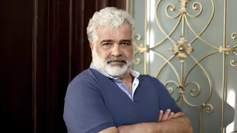 In questa immagine, si vede un uomo maturo con capelli bianchi e una barba folta. Indossa una maglietta blu e ha un'espressione seria, con le braccia incrociate. Sullo sfondo, ci sono elementi decorativi di metallo che aggiungono un tocco elegante all'ambiente. La luce mette in risalto i dettagli del suo viso e dell'abbigliamento.