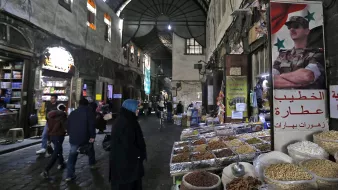 L'image montre une scène d'un souk ou d'un marché typique, probablement en Syrie. On aperçoit des étals avec des épices, des fruits secs, et d'autres produits en vente. Les murs de la rue sont en pierre et semblent anciens, créant une ambiance historique. Des personnes, dont certaines portent des vêtements traditionnels, se déplacent dans la rue, tandis qu'une affiche avec un symbole militaire est accrochée à un mur, ajoutant un élément de contexte culturel et politique à la scène. La lumière faible et les ombres accentuent l'ambiance conviviale et animée du marché.
