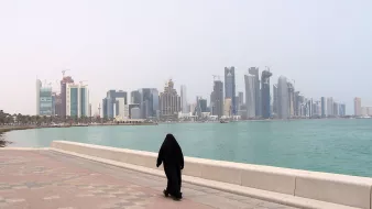 L'image montre une personne vêtue d'une tenue traditionnelle noire, marchant le long d'une promenade au bord de l'eau. En arrière-plan, on aperçoit une ligne d'horizon urbaine avec des gratte-ciel modernes et des bâtiments en construction, représentant une ville côtière. L'ambiance est calme, avec un ciel légèrement brumeux et une mer tranquille. Les pavés de la promenade ajoutent une touche de couleur à la scène.