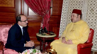 L'image montre deux hommes assis dans une pièce richement décorée. L'un est vêtu d'un habit traditionnel jaune et porte un fez, tandis que l'autre est en costume. Ils sont engagés dans une conversation, avec des expressions sérieuses. En arrière-plan, on peut voir des éléments décoratifs, tels que des rideaux rouges et un arrangement floral sur une table.
