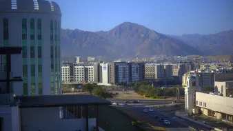 L'image montre un paysage urbain avec des bâtiments modernes à l'avant-plan, surmontés par une vue de montagnes majestueuses à l'arrière-plan. Le ciel est clair et bleu, ce qui donne une belle luminosité à la scène. En bas, on aperçoit une route avec des voitures, et les bâtiments montrent une architecture contemporaine, témoignant d'une ville dynamique et en développement. Les montagnes, quant à elles, ajoutent une dimension naturelle impressionnante au paysage urbain.
