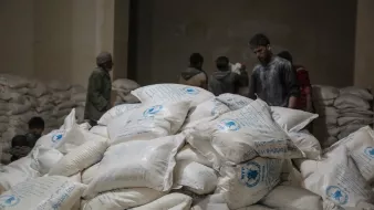 تظهر في الصورة مجموعة من الأشخاص داخل مكان يبدو أنه مخزن للمواد الإغاثية. في المقدمة، هناك كومة كبيرة من الأكياس البيضاء التي تحمل علامات معينة، ويبدو أن الأكياس تحتوي على مواد غذائية أو إغاثية. في الخلفية، يمكن رؤية بعض الأشخاص الذين يتحدثون أو يعملون. الإضاءة في المكان خافتة، مما يعطي شعورًا بالجدية والاهتمام بالعمل الذي يقومون به.